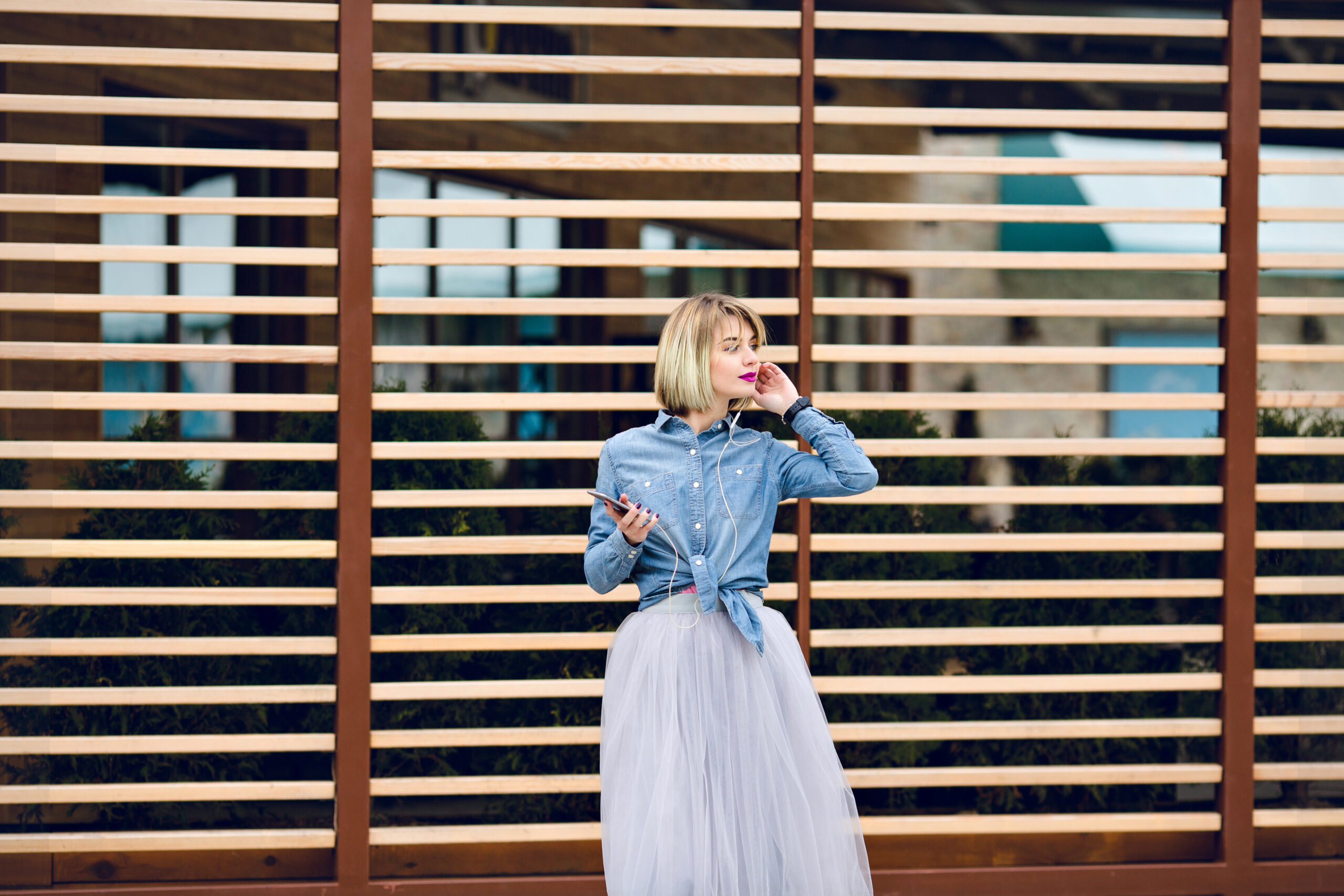 Dreamy blonde girl with bright pink lips standing and listening to music on a smartphone with striped wooden balks behind wearing blue jeans shirt, grey tulle skirt and black sneakers.
